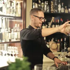 Barman w okularach nalewa drinka do kieliszka. Za nim półki z butelkami alkoholu.