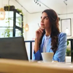 Kobieta w niebieskiej marynarce siedzi przy biurku z laptopem i kawą, zamyślona.