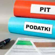 Dwa segregatory, jeden czerwony z napisem PIT, drugi niebieski z napisem PODATKI, leżą obok formularzy podatkowych i długopisu.