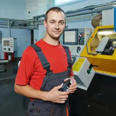 Uśmiechnięty tokarz w warsztacie trzyma przyrząd pomiarowy. W tle maszyna CNC i inne urządzenia.