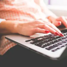 Dłonie z czerwonymi paznokciami piszą na laptopie. To idealny obrazek, gdy szukasz copywriter praca.