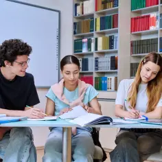 Trzech studentów przy biurku, otoczonych książkami, pogłębia swoją wiedzę. Ich zaangażowanie w naukę to klucz do przyszłego wykształcenia.