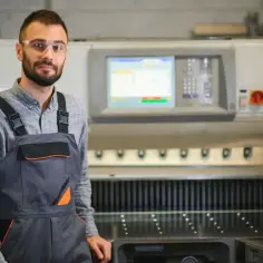 Młody operator CNC w okularach ochronnych i ogrodniczkach stoi przed maszyną.