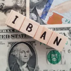 Drewniane klocki z napisem IBAN leżą na banknotach dolarowych. IBAN to międzynarodowy numer rachunku bankowego.