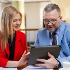 Kobieta w czerwonej marynarce i mężczyzna w niebieskiej analizują dane na tablecie, omawiając strategię biznesową. Ich wspólna praca nad projektem MBA przynosi efekty.