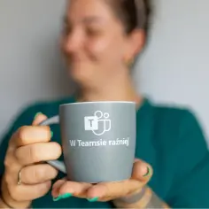Kobieta trzyma kubek z logo Microsoft Teams i napisem "W Teamsie raźniej". Idealny do kawy podczas pracy zdalnej lub rozmów z helpdeskiem.