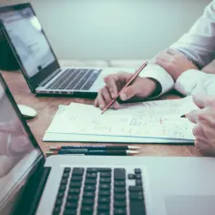 Dwóch mężczyzn pracuje nad dokumentami, jeden z nich pisze, drugi wskazuje palcem. Na stole dwa laptopy, długopisy i notatki. Może to być analiza danych, planowanie projektu lub sprawdzanie numeru NIP.