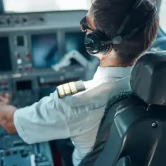 Pilot samolotu w słuchawkach i okularach przeciwsłonecznych, z ręką na sterach, skupiony na locie.