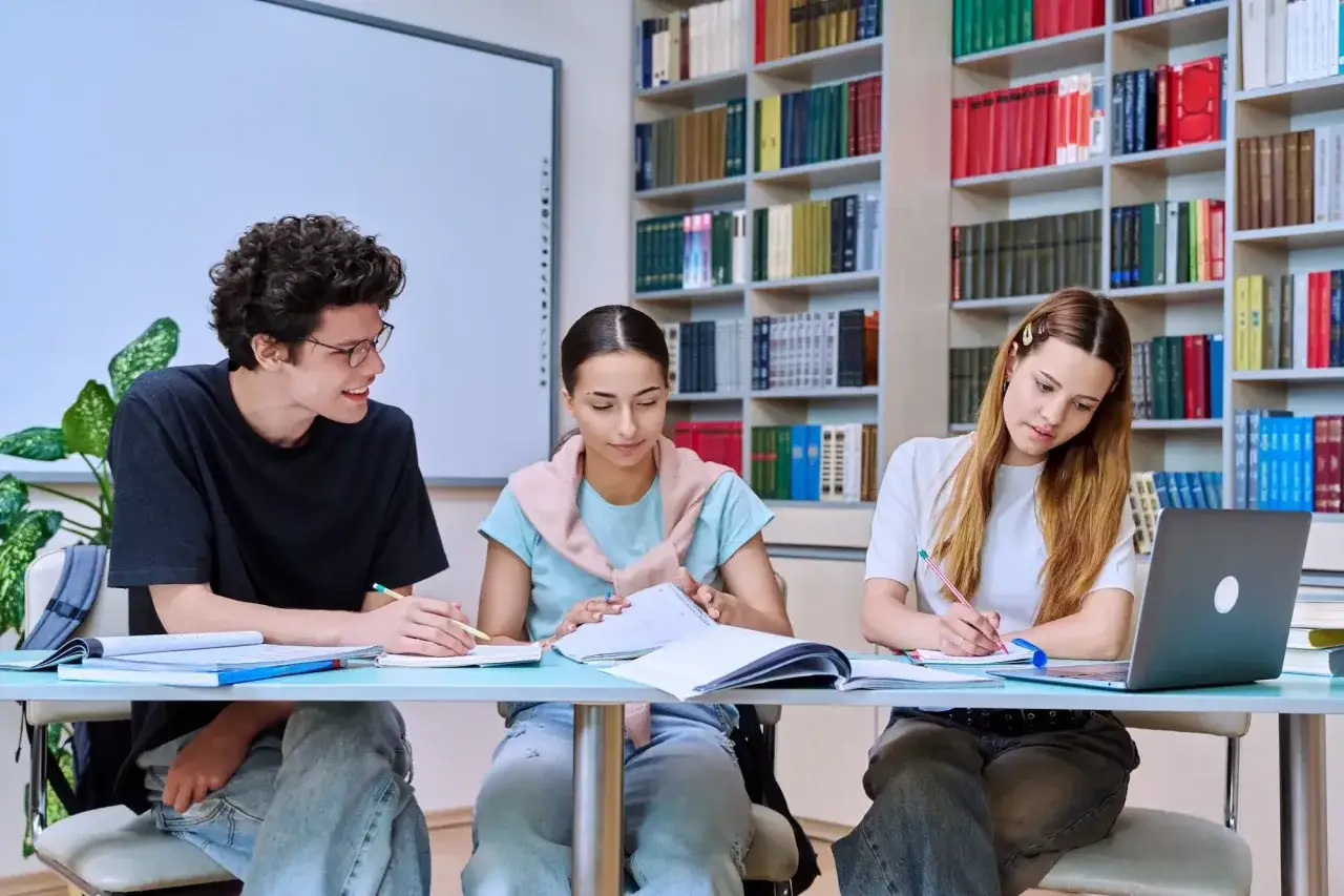 Trzech studentów przy biurku w bibliotece, otoczonych książkami, skupionych na nauce. Ich zaangażowanie świadczy o dążeniu do zdobycia wykształcenia.