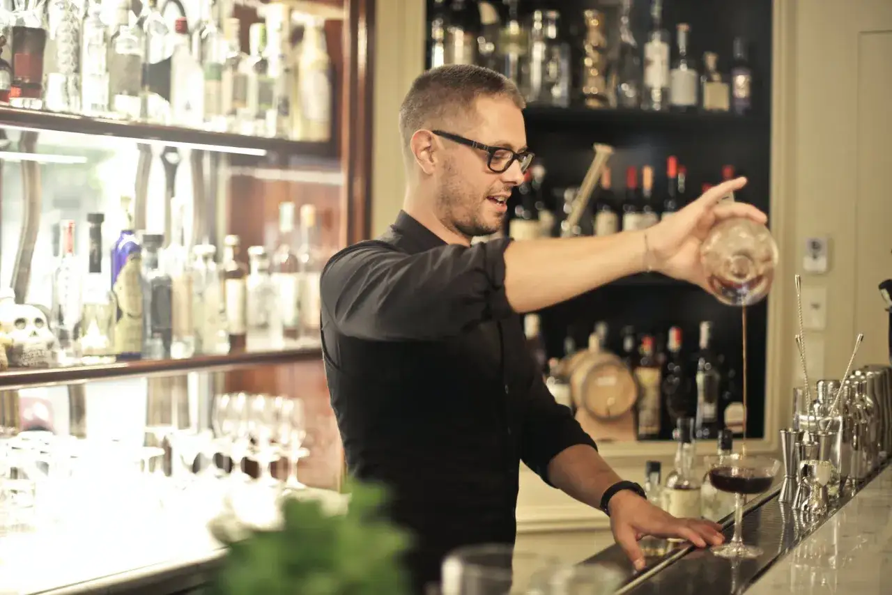 Barman w okularach nalewa drinka do kieliszka. Za nim półki z butelkami alkoholu.