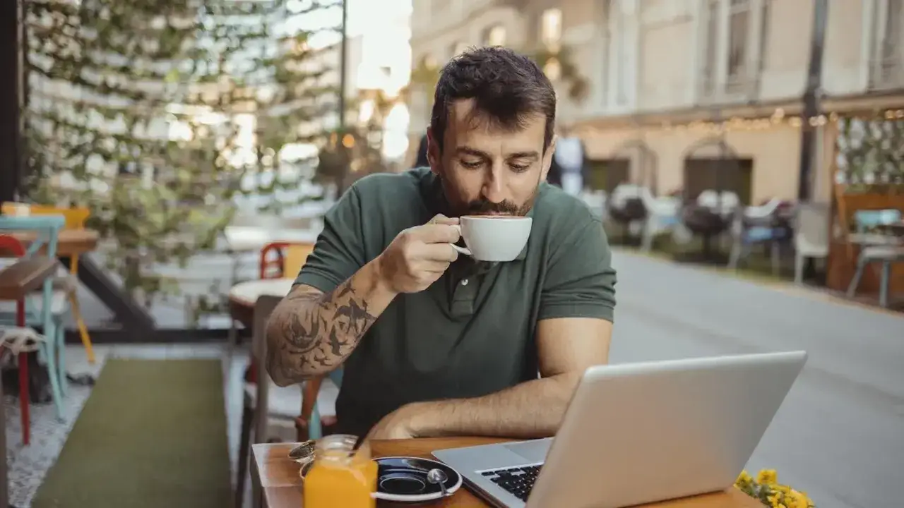 Dziennikarz z tatuażami na ramieniu popija kawę, pracując na laptopie w kawiarnianym ogródku.
