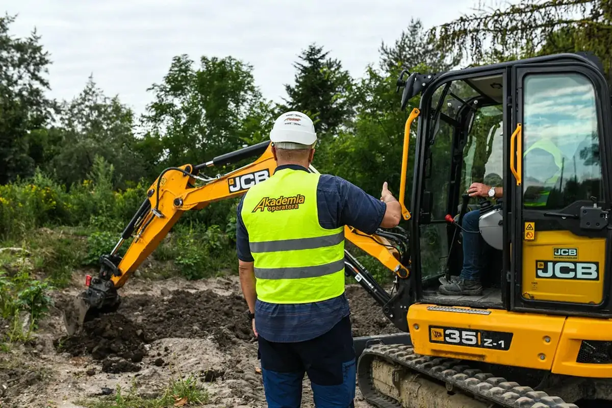 Operator koparki w kamizelce odblaskowej instruuje kolegę w kabinie maszyny.