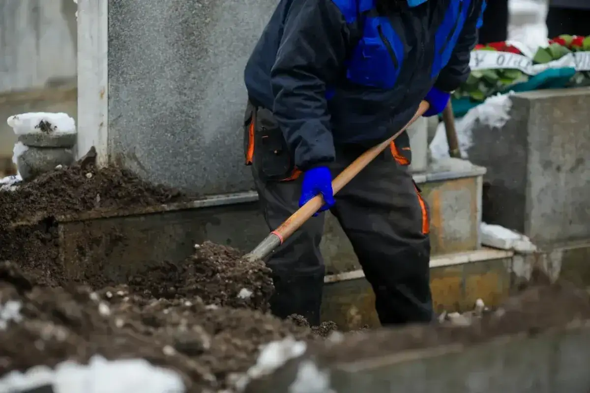 Grabarz w niebiesko-czarnej kurtce i pomarańczowych spodniach kopie łopatą ziemię przy nagrobku.