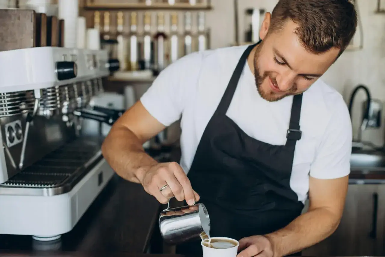 Uśmiechnięty barista w czarnym fartuchu nalewa mleko do kawy przy ekspresie.