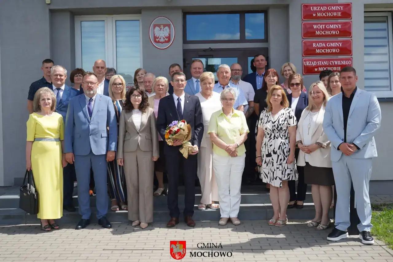 Grupa osób przed urzędem gminy. Wójt gminy Mochowo i radni pozują do zdjęcia. Ciekawe, ile zarabia wójt.