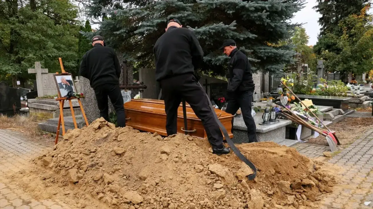 Grabarze przygotowują grób. Na pierwszym planie stos ziemi, obok trumna i portret zmarłego.